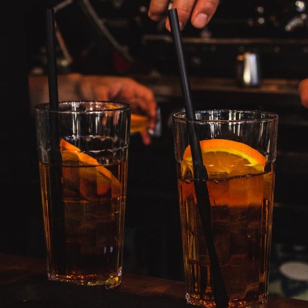 Two tall glasses of amber drinks with orange slices and black straws on a dark bar.